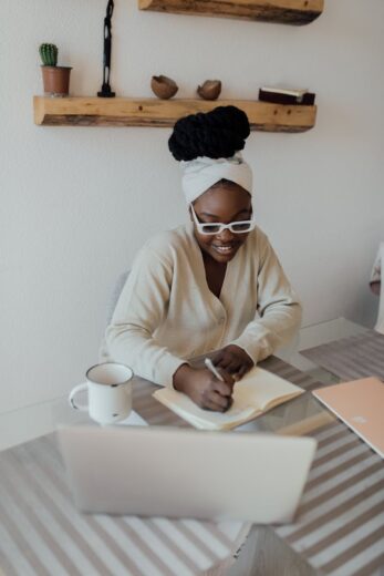 African American woman sitting indoors writing in a journal at a desk with a laptop and coffee mug.